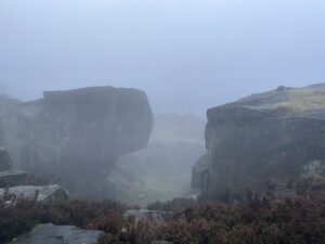 A rugged moorland landscape. There is a big overhanging cliff. It is very foggy.